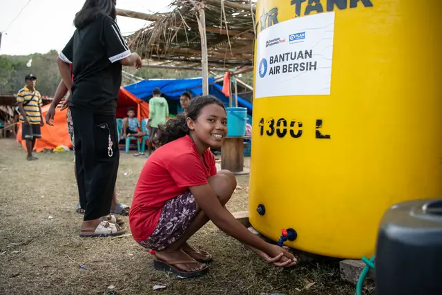 A girl smiles while rinsing her hands at the tap of a 1,300-litre aid water tank